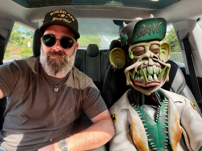 A bearded man wearing sunglasses and a 'Bored Ape Yacht Club' cap sits in a car next to a Bored Ape character figure wearing a matching BAYC cap.