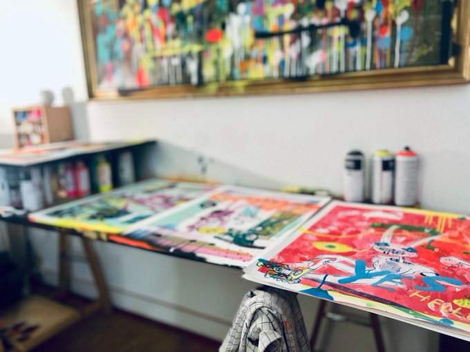 An artist's studio workspace featuring several colorful spray-paint canvases laid out on tables, with a row of spray paint cans lined up against a white wall beneath a large, framed abstract painting.