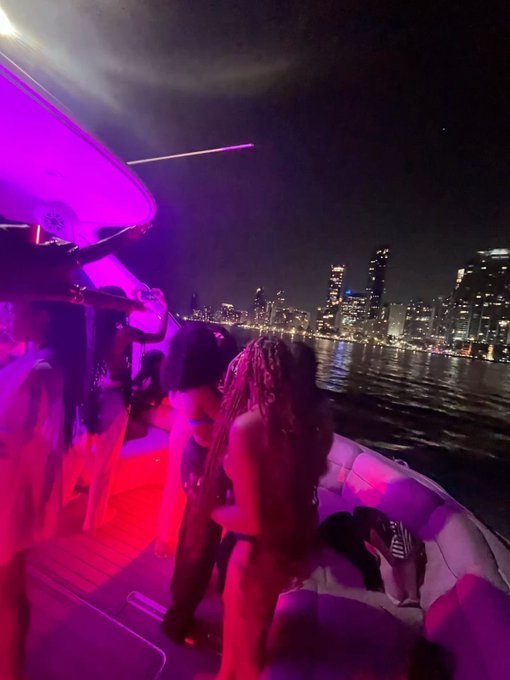 A vibrant night scene on a yacht with people dancing under pink and purple lighting, overlooking a city skyline across the water, captioned "GM" likely referencing "Good Morning" in hip-hop/club culture.