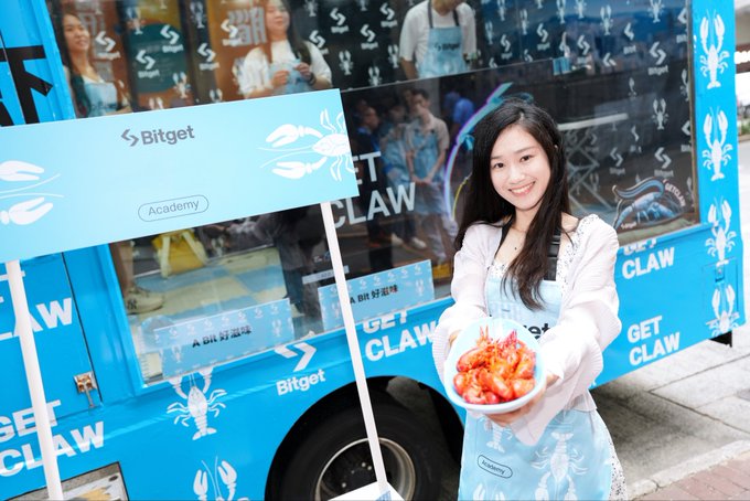 A smiling young woman in a Bitget-branded apron holds a plate of cooked crawfish in front of a blue "Bitget" truck promoting a "Get Claw" event at MRT Yuanshan Station for TaipeiWeek.