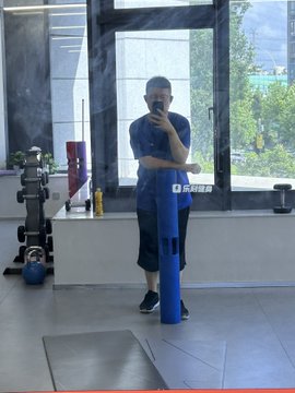A mirror selfie of a man holding a blue foam roller, wearing a black outfit with Chinese characters '苏州联胜' (Suzhou Liansheng) on it, standing in a room with large windows.
