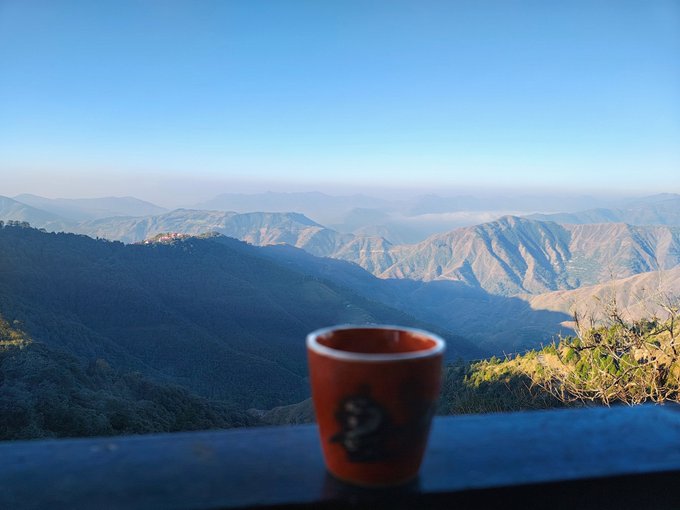 A red coffee mug sits on a railing overlooking rolling mountain ranges under a clear blue sky, capturing a serene Sunday morning. The image embodies the post's message of taking time to recharge and enjoy nature's beauty rather than grinding nonstop.