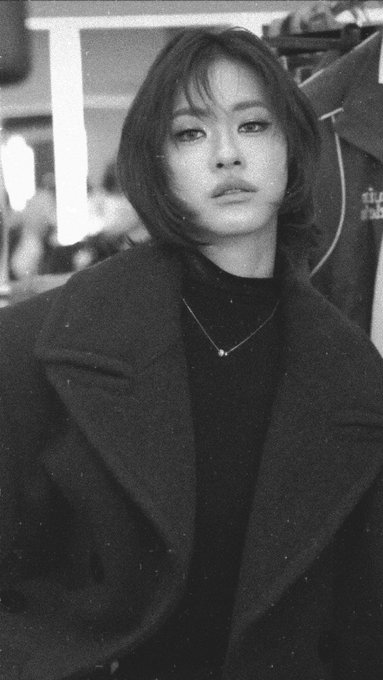 A black-and-white portrait of a young woman with a short bob haircut, wearing a dark coat and turtleneck with a delicate necklace.
