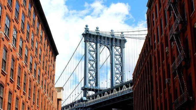 A view of the Manhattan Bridge tower framed between two red brick buildings against a cloudy blue sky, identical to the first image.