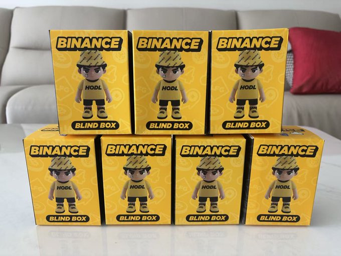 Seven yellow "BINANCE BLIND BOX" packages featuring a cartoon figure wearing a "HODL" shirt and a patterned hat are stacked in two rows on a white table, matching the post about collecting a complete set of Binance's "Palu" blind boxes.