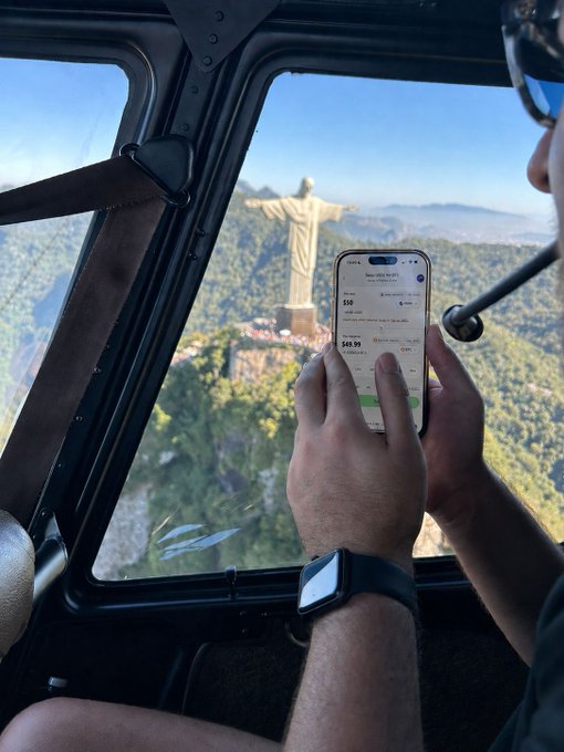A passenger on a helicopter holds a smartphone displaying a crypto trading app ($59, $48.98 prices visible) while approaching the iconic Christ the Redeemer statue in Rio de Janeiro, illustrating the post's message that crypto markets are accessible anywhere—even 1,000 feet above a world wonder.