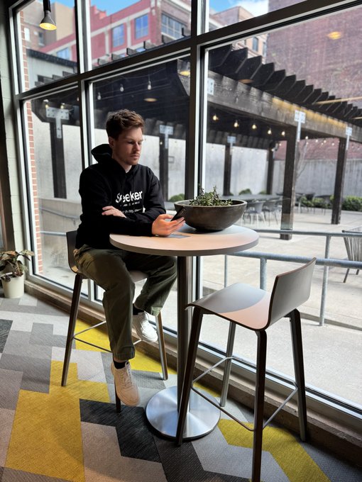 The image shows a man sitting alone at a table with his arms crossed while looking out of an open window. The scene takes place in front of a building, possibly a restaurant or cafe. There are two chairs visible near the table where the man is seated - one chair on each side. A potted plant can be seen placed next to the man's seat, and there is also a bowl with some plants sitting on top of it. The image seems to be related to a post about "Happy Morning Seeker gang New week. Time to attack your rings. Make sure to stake a small amount of $SKR at least a few times daily, use some dapps, do on chain swaps."