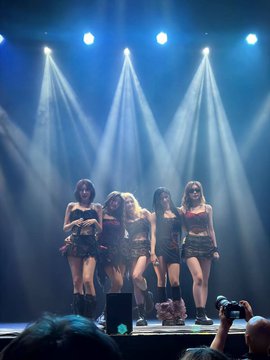 A group of performers in matching dark outfits dance on a stage illuminated by dramatic blue spotlights, with audience members visible in the foreground.