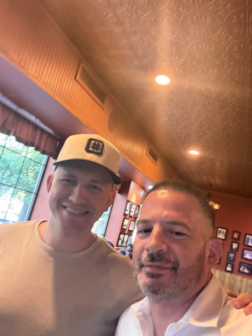 Two men pose together for a selfie in a restaurant or diner, one wearing a beige cap and t-shirt, the other in a white shirt with a short beard.