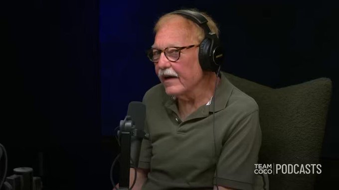 An older man with glasses and a mustache wearing headphones and a green polo shirt speaking into a microphone on a Team Coco Podcasts set.