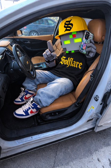 A digitally edited photo of a person in a car wearing a yellow cap, a "Solflare" hoodie, and red/white/blue sneakers, with a Minecraft-style creeper face and blocky hands holding a phone.