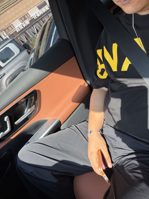 A man in a black t-shirt with yellow lettering sits in a tan leather car seat, holding a phone with a brown case, while wearing a seatbelt. The image shows a passenger enjoying a ride, tying into the celebratory mood as the caption mentions a birthday celebration and friends gathering together.