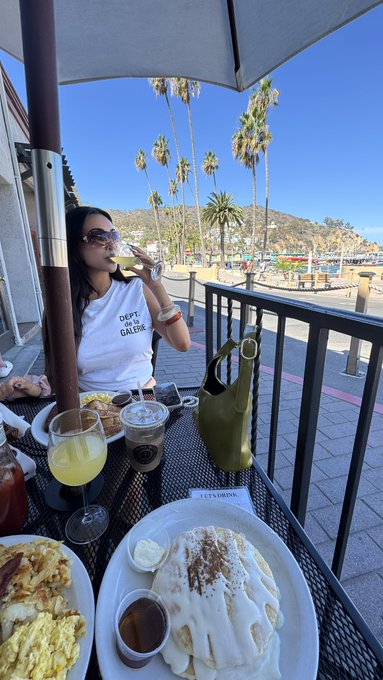 A woman wearing sunglasses and a 'DEPT OF FASHION GALORE' t-shirt sits at an outdoor patio table with food, a drink, and a view of palm trees and hills.