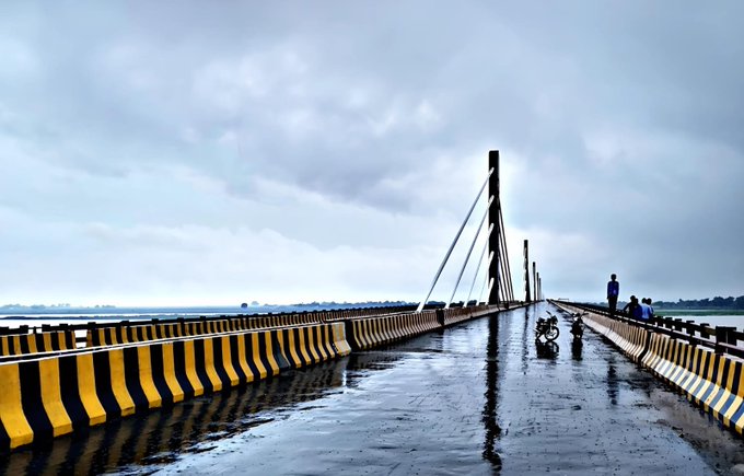 A wet bridge over water under a cloudy sky, with a motorcyclist and pedestrians visible in the distance and yellow-and-black striped barriers along the sides.