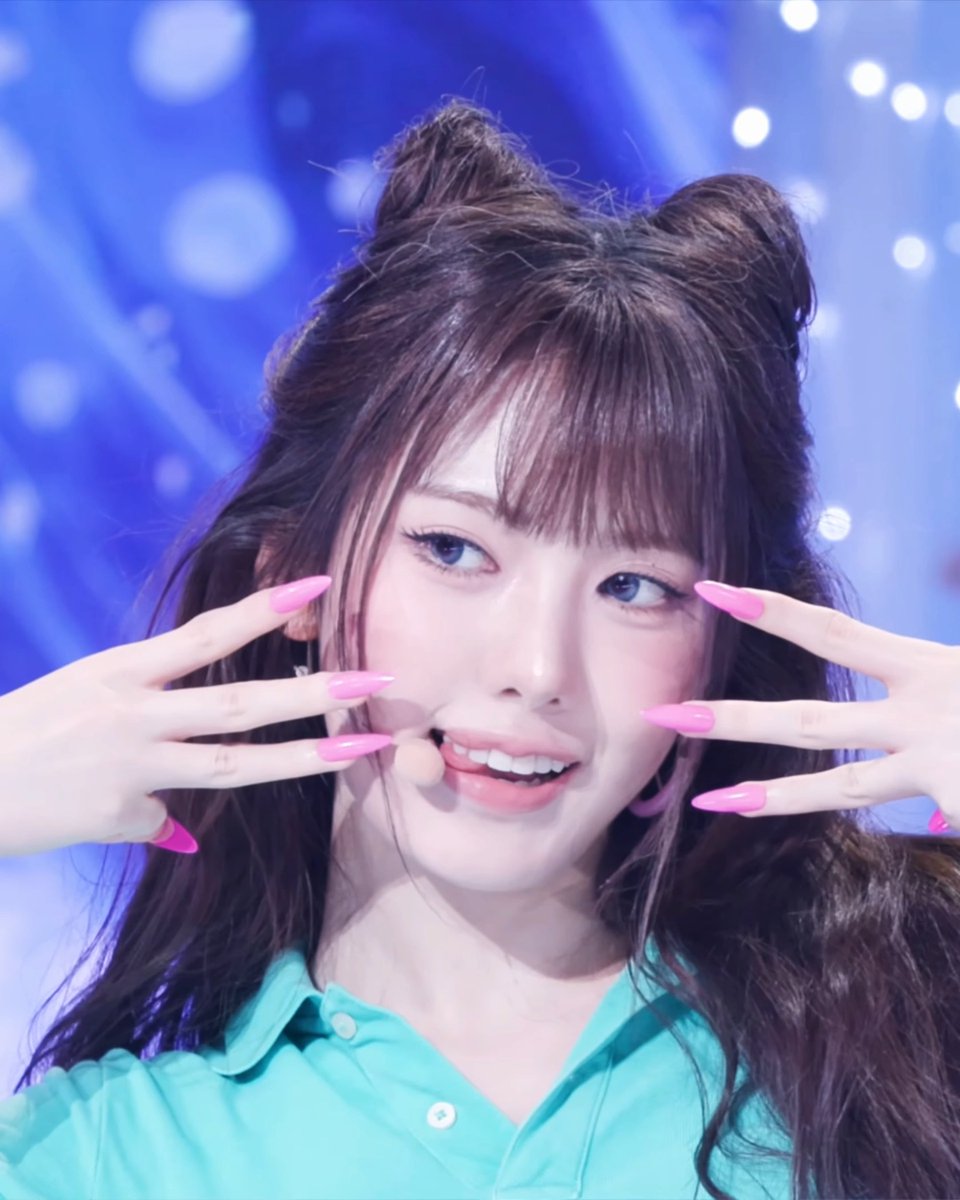 A young woman with long brown hair styled in space buns and pink manicured nails poses playfully with her hands framing her face against a blue, sparkly background while wearing a turquoise button-up shirt.