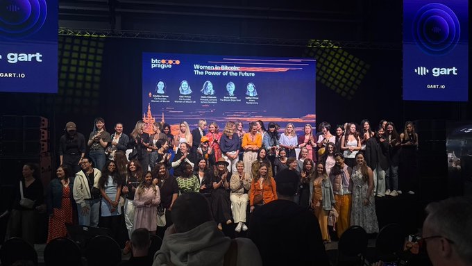 The image shows a large group of people gathered together, with some standing and others sitting, posing for a photo. The crowd appears to be composed of both men and women, all dressed similarly in black clothing. They are positioned in front of a stage that has a screen displaying an event or presentation related to Bitcoin. The text "Women Innovators of the Future" is visible on the screen, indicating that this gathering may be part of a Women's Innovation Summit.