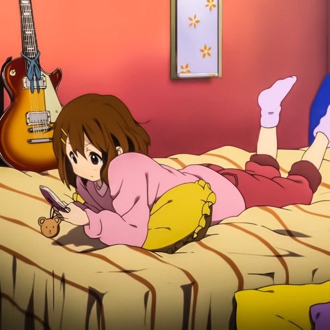 An anime scene showing a girl with brown hair lying on a bed and looking at a pink flip phone, with a guitar leaning against the wall behind her.