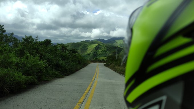 A scenic mountain road shot from behind a motorcyclist wearing a bright green helmet, with lush green hills and cloudy skies in the distance.