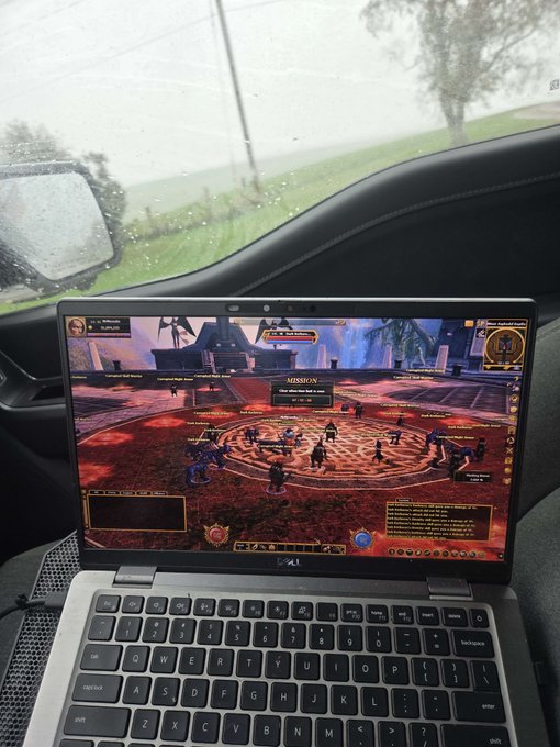 A laptop displaying a fantasy MMORPG game is set up on the passenger seat of a car, with a rainy view visible through the windshield.