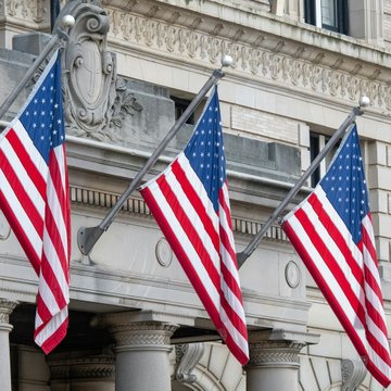 Three American flags hang from the ornate stone facade of a classical-style building, symbolizing U.S. national interests. The image visually anchors the post's headline about the United States becoming a net Bitcoin importer for the first time since 2022, with large transfers exceeding $10 million driving the shift in digital asset flows as of June 2026.
