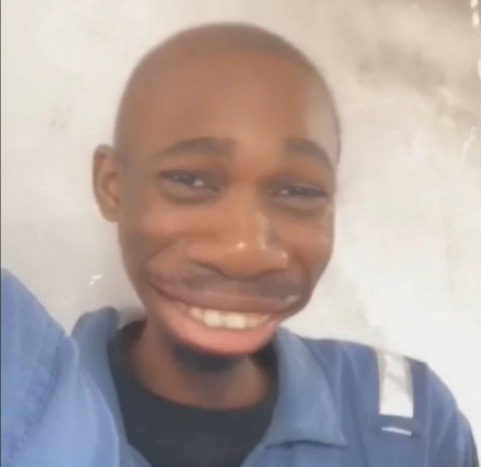 A close-up selfie-style photo of a smiling young Black man with a shaved head, wearing a blue work shirt with a reflective stripe.