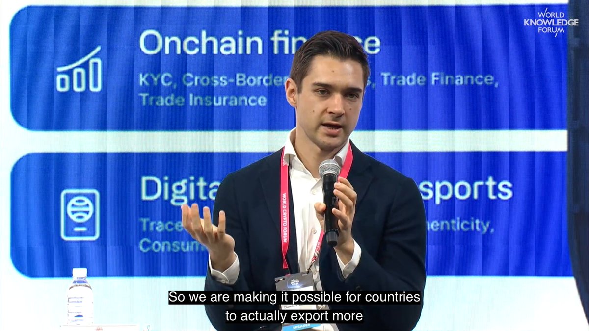 The image shows a man standing in front of a blue screen with white text, giving him a podium-like appearance as if he's speaking at an event or conference. The background also features other text elements such as "Onchain" and "Focus on". A microphone is attached to the man, indicating that his speech is likely being amplified for better visibility in the room. 

The main focus of the image is the man who appears to be delivering a presentation or speech, and it conveys a professional context with the presence of a conference or event setting. The man's stance and use of the microphone suggest that he wants his audience to hear him clearly, emphasizing the importance of effective communication in such environments.
