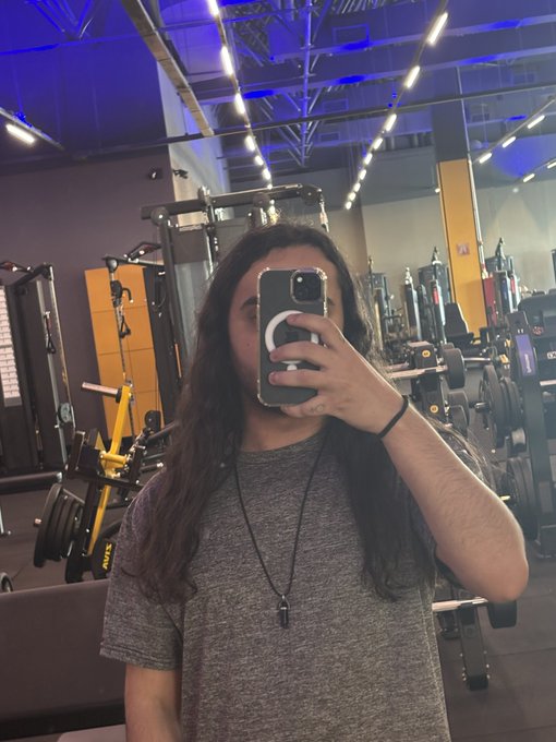 A mirror selfie of a person with long dark hair holding a smartphone in a gym setting with weightlifting equipment and blue lighting visible.