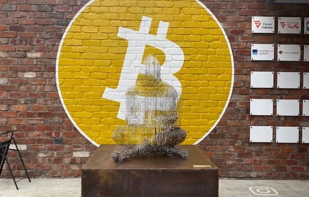 An indoor shot of an art installation featuring a wireframe sculpture sitting on a pedestal in front of a brick wall painted with a large yellow Bitcoin logo and Bitcoin logos on adjacent panels.