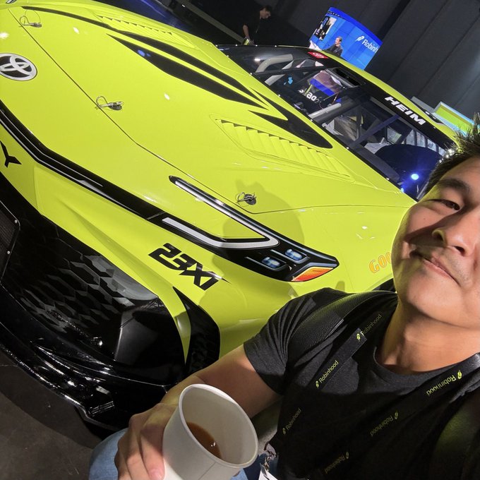 A man in a black lanyard takes a selfie holding a cup in front of a bright neon yellow sports car marked '25X1' and 'HEIMA' at an indoor exhibition.