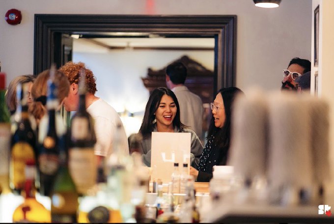 Two women laughing together at what appears to be a bar or event, with out-of-focus bottles in the foreground.