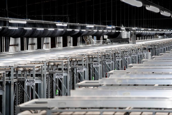 Rows of industrial Bitcoin mining rigs (labeled A015–A023) hum in a data center, visually representing the large-scale mining operation behind Eric Trump's "American Bitcoin" company and its HODL of over 8,000 $BTC.