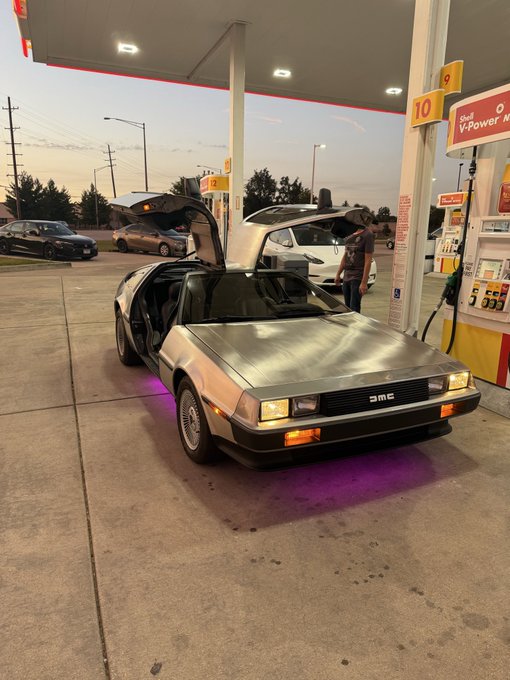 A DeLorean DMC-12—iconic for its gull-wing doors and stainless steel body—sits at a Shell V-Power gas pump, with the "10" and "9" fuel grade signs visible overhead. The "Back to the Future" time machine serves as a witty metaphor for the post's message: just as Doc Brown's DeLorean represents a vehicle that defies time, the "$Laura" token is presented as a "time-traveling" investment that early investors will miss out on once its value becomes widely known.
