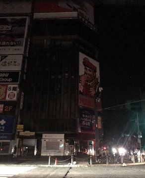 A nighttime street photo in Japan showing a building corner with various illuminated signs and billboards, including 'UNIQLO' and '2F', with pedestrians and cyclists visible on the lit street below.