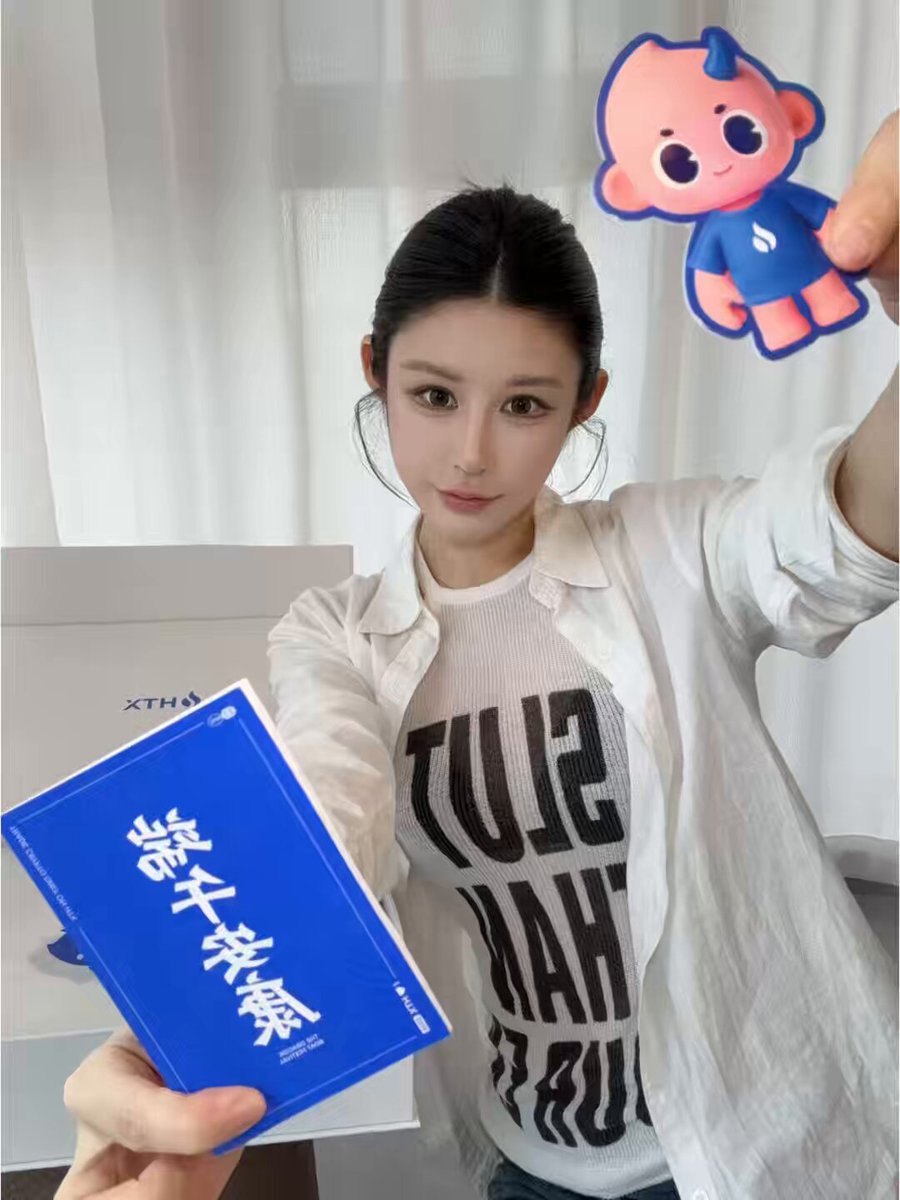 The image features a woman holding up two items, one of which appears to be a book with Asian characters written on it. The other item is a small toy or figurine that resembles a baby. The woman seems to be posing for the photo while holding these objects. The background of the image shows a window and a chair, suggesting that she might be in her home or office setting.
