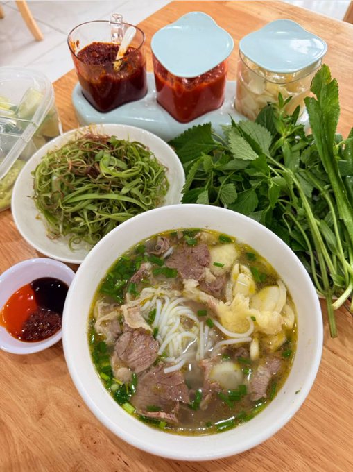 A bowl of Vietnamese pho with beef and noodles is served alongside fresh herbs, bean sprouts, chili sauce, hoisin sauce, and other condiments on a wooden table.