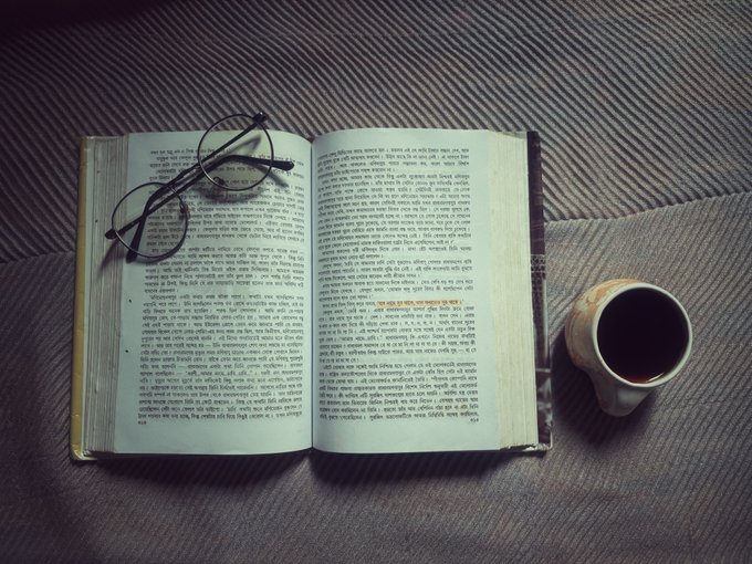An open book with text in what appears to be Bengali or a similar script lies on a striped fabric surface, with a pair of black eyeglasses resting on the left page.