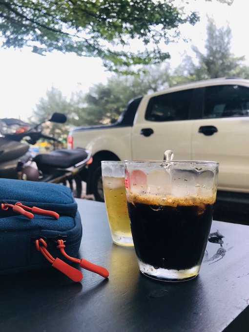 Two glasses of cold beverages (one appears to be dark beer and the other a lighter drink) sit on a black table next to a blue bag with orange zippers, with a white pickup truck and motorcycle visible in the background.