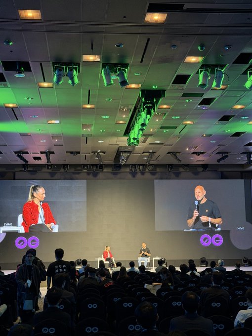 A large conference hall where two speakers are seated on stage, displayed on big screens to an audience. The event appears to be a tech or industry talk, with green stage lighting and purple "oo" logos visible.