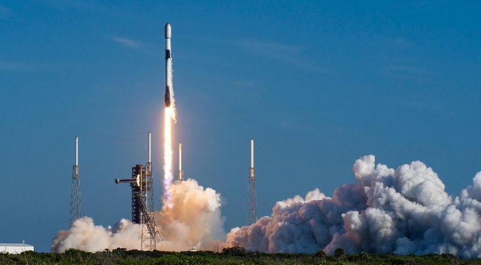 A SpaceX Falcon 9 rocket launches into a blue sky, emitting bright flames and a large plume of smoke from the launch pad.