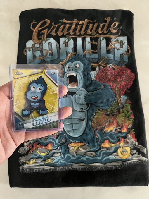 A hand holding a 'GRATITUDE GORILLA' trading card in front of a black plush blanket featuring matching gorilla artwork.