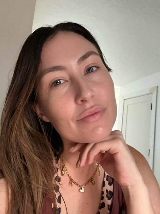 A close-up selfie of a woman with long brown hair, wearing a leopard print top and gold layered necklaces, with a small mole visible on her cheek.
