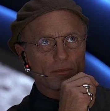 A close-up of an older man wearing a brown cap, round wire-rimmed glasses, and a headset with a microphone, looking contemplative with his hand near his chin, showing silver rings.