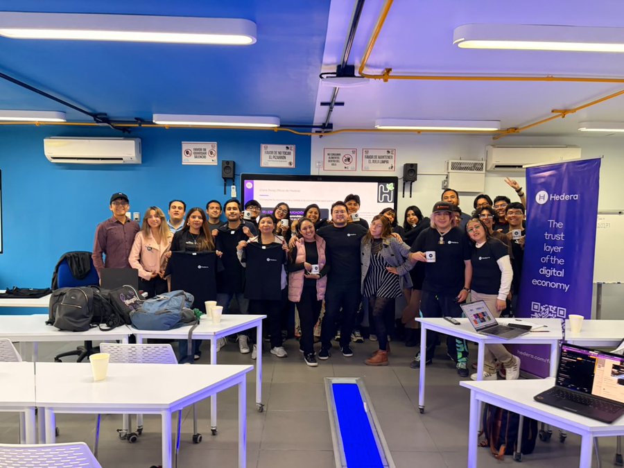 A group of around 25 people pose together at the first Hedera meetup at UNAM (Universidad Nacional Autónoma de México), with a banner reading "Hedera - The trust layer of the digital economy" visible on the right side of the room. The attendees, many wearing Hedera-branded shirts, are gathered in a classroom-style space with laptops and educational materials, reflecting the post's focus on bringing Hedera education and ecosystem training to university students in Mexico.