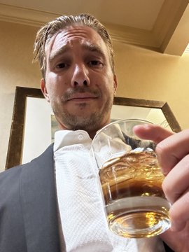 A man with a goatee, wearing a white shirt and dark jacket, holds up a glass of an amber beverage (likely whiskey) towards the camera in a selfie-style shot.
