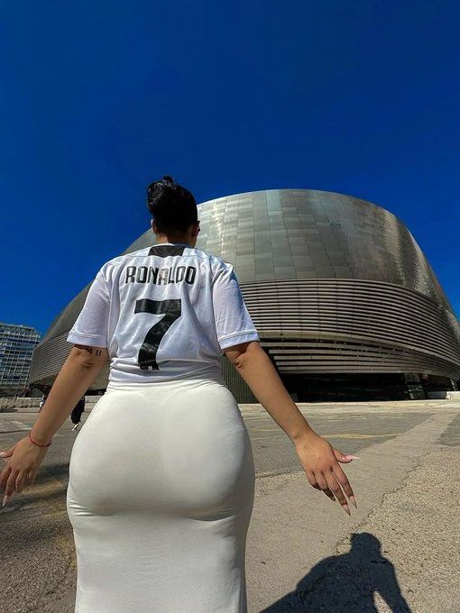 A photo of a woman from behind wearing a white soccer jersey with 'RONALDO' and the number 7, and a tight white skirt, standing in front of a modern metallic dome-shaped building under a clear blue sky.