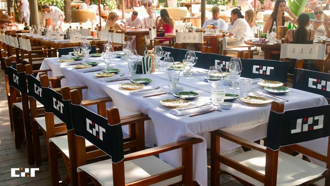 An outdoor dining scene with long tables set with white tablecloths, fine glassware, and chairs branded with 'COT' logos, surrounded by patrons in a sunny garden setting.