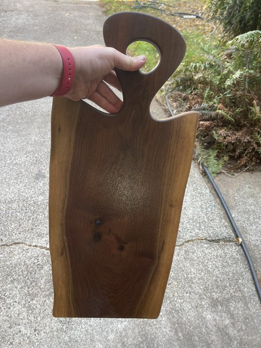 A hand holding a large, dark wooden cutting or serving board with a thumb hole and wavy live edges, photographed outdoors on a concrete surface.