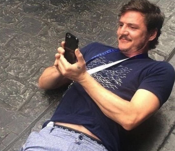 A photograph of actor Pedro Pascal lying back on a cobblestone surface, smiling broadly while looking at a black smartphone he is holding in his hands.