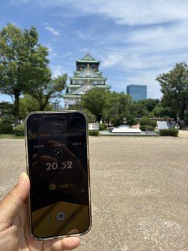 A hand holds a smartphone in front of Osaka Castle, displaying a fitness app reading of "20.32" with a "+168.63" gain, likely from a STEPN-related walking session. The image ties to the post about reaching Osaka Castle, with the phone showing nearly half the user's daily step goal achieved.