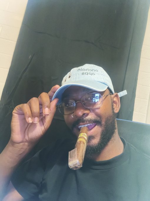 A man wearing glasses and a light blue 'Original 2020' cap tips his hat while smiling with a cigar in his mouth.